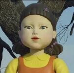 图片中是一个造型可爱的小女孩玩偶，她有着棕色的头发并扎成了双马尾，上身穿着黄色的上衣，搭配橙色的背带裙。背景是模糊的树枝和天空，整体画面简洁，玩偶形象显得乖巧温馨。