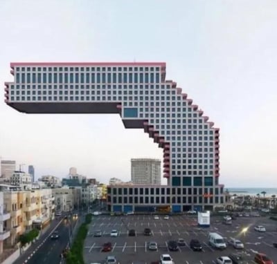 这是一张经过创意合成的海滨城市建筑景观图，画面中的高层建筑被修改成违背物理常识的形态：建筑下半部分正常坐落于地面，上半部分却大幅弯折，末端完全悬浮在空中，形成一个类似“拱门”的奇特造型。画面下方是城市道路与露天停车场，能看到行驶和停放的车辆，背景是错落的城市楼宇与远处的海洋，整体场景充满超现实的荒诞感，打破了人们对常规建筑的认知，带来强烈的视觉冲击。