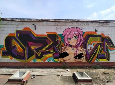 一幅街头涂鸦作品，展示了一位粉色头发、绿色眼睛的动漫风格少女，她有着惊讶的表情，双手举在胸前。少女形象被黑色 censorship bars 遮挡胸部。背景是色彩鲜艳的涂鸦文字，主要使用紫色、橙色、黄色和黑色。整个作品绘制在一面白色砖墙上，墙面上有一些剥落和损坏的痕迹。墙下有一些混凝土结构和碎片，背景是晴朗的蓝天和少量白云。