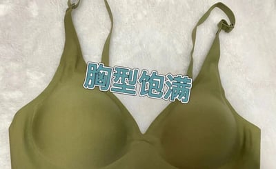 图片中展示了一件绿色的内衣，放置在白色毛绒质感的背景上。内衣整体为简约设计，肩带可调节，杯型呈现自然弧度。图片中央有一行蓝底白边的文字“胸型饱满”，直观突出该内衣在塑造胸型方面的特点，整体风格偏向产品展示，旨在宣传内衣的塑形效果。