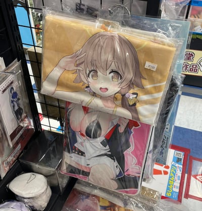 动漫周边商店货架上展示的两款角色商品，上方是黄色背景的毛巾，印有棕色短发、黄色眼睛的动漫女孩敬礼图案，价格标签显示¥2180；下方是粉色背景的抱枕，印有穿着性感的动漫女性角色。周围可见其他动漫周边产品，整体呈现典型的二次元商品陈列场景。