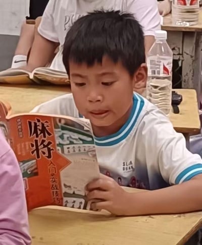 课堂场景中，一名穿着白色校服的小学生正专注地阅读一本橙色封面的《麻将入门实战技巧》，与周围同学阅读课本的场景形成鲜明反差。男孩表情认真，仿佛在钻研重要教材，而桌上的矿泉水瓶和背后同学的课本更凸显了这一画面的荒诞幽默感。