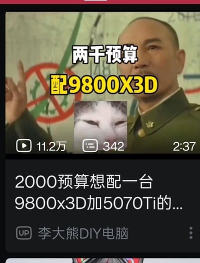这是一张B站数码类视频的截图，画面上半部分使用了抗战剧《亮剑》中楚云飞的剧照，角色抬起右手食指指向上方，搭配白色文字“两千预算”和黄色醒目文字“配9800X3D”，文字下方叠加了一张眯眼憋笑的猫咪表情包，暗示该需求的不合理与荒谬。画面下方是B站视频的信息栏，显示该视频播放量为11.2万，评论数342，时长2分37秒，视频标题为“2000预算想配一台9800x3D加5070Ti的...”，UP主为“李大熊DIY电脑”，整体内容是调侃DIY电脑领域部分消费者提出的远低于市场正常价格的不合理配置需求的搞笑玩梗内容。