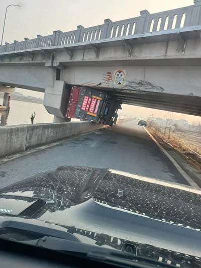 图片展示了一辆红色货运卡车卡在混凝土立交桥桥洞下的惊险场景。卡车车身严重倾斜，前半部分（含车厢）被桥体底部卡住，无法移动，车厢侧面可见红色文字标识（内容不清晰）。照片拍摄视角来自一辆行驶中的汽车内部，下方清晰可见汽车的仪表盘和布满灰尘的挡风玻璃。桥体为灰色混凝土结构，带有白色护栏，桥洞上方有模糊的警示标识。桥下道路为双向车道，远处有一辆黄色轿车正常行驶。左侧可见河流及岸边光秃树木，右侧为荒地，整体环境光线柔和，推测为清晨或傍晚时分。