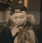 图片中是1986版电视剧《西游记》中的经典角色猪八戒，由演员马德华饰演。他头戴黑色帽子，帽上有金色装饰，脖子围着棕色围巾，右手自然地放在嘴边，面部表情滑稽搞怪，眼神中带着一丝狡黠，整体呈现出轻松诙谐的氛围。这一截图常被网友用作表情包，表达搞笑、调侃或略带顽皮的情绪。