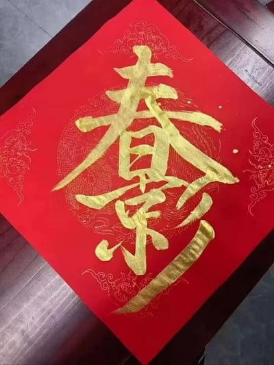 这是一张充满新春氛围的书法作品，以带有精致龙纹暗花的红色宣纸为底，用金墨书写了将“春”与“影”结合而成的合体字，字体苍劲有力，金墨在红底上格外醒目，整体风格喜庆红火，是春节期间常见的民俗书法创作，可用于家居装饰或新春相关场景，传递新春的美好氛围。