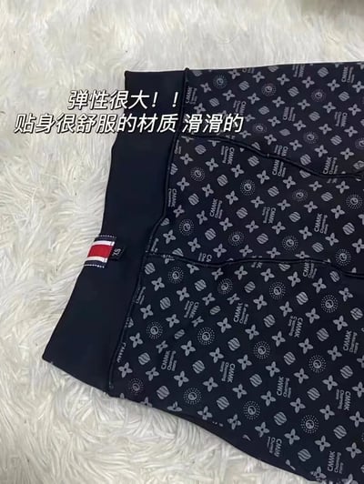 图片展示了一件深色底带有灰色花纹图案的服饰，上面印有重复的花朵、圆圈图案和"CMNK"文字。服饰的腰部有深蓝色松紧带设计，带有一个红白蓝三色条纹的标签，上面有"SK"字样。图片左上角有文字标注："弹性很大！！贴身很舒服的材质 滑滑的"，表明该服饰具有弹性好、材质舒适顺滑的特点。服饰被放置在白色毛绒质地的背景上，整体展示了产品的材质和图案细节。