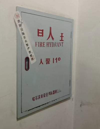 一张实拍的消防栓箱照片，箱体为灰色，上面有红色的中文标识和英文"FIRE HYDRANT"字样。最显眼的是中文标识出现明显错别字，本应是"消火栓"的位置写成了"日人 王"，下方还有"人警 110"的错误标识。左侧贴有一张竖条标签，写着"消防设施 非火警严禁使用"并盖有红色印章。底部有小字显示生产厂家信息"哈尔滨市荣达消防器材工"。这张图片因为明显的错别字而显得十分搞笑，可能是生产厂家在制作标识时出现的失误。