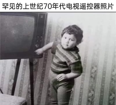 这是一张带有幽默玩梗性质的内容图，顶部配有文字“罕见的上世纪70年代电视遥控器照片”，图中是一张上世纪70年代的原始家庭老照片，画面里一个穿着条纹短袖上衣和紧身裤的卷发幼儿，正伸手操作老式立式电视机的按键，身体微微前倾，另一只手放在腰侧。这张图的笑点在于用玩梗的标题将手动帮忙操作电视的幼儿调侃为那个年代的“人肉遥控器”，因为早期的电视机没有无线遥控器，只能手动按键操作，通过这种反差感制造出怀旧又搞笑的效果，既引发经历过那个时代的人的共鸣，也能让当代人感受到家电发展的时代变化。