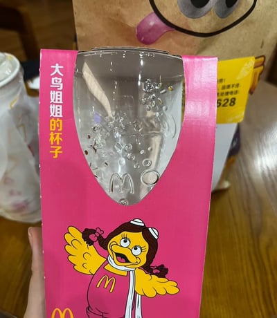 图片展示了一个麦当劳的粉色包装盒子，里面装着一个透明的杯子，杯身上有麦当劳标志。包装上印有一个卡通形象的鸟姐姐，她有着棕色的头发扎着双马尾，穿着带有麦当劳标志的粉色衣服，张开双臂做出欢迎的姿势。包装左上角有竖排文字'大鸟姐姐的杯子'。背景中可以看到一个带有卡通脸设计的棕色纸袋和部分白色杯子。