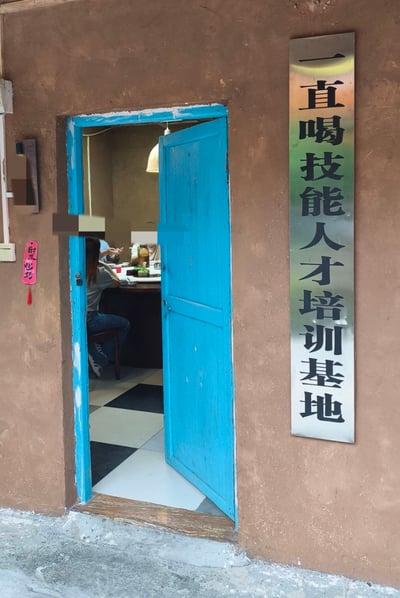 这是一张线下小店门口的实拍照片，建筑外墙为土棕色质感，蓝色的木质单开门半开向内，门内可见黑白格子地砖，还有数人围坐在圆桌旁用餐，能看到桌上的餐食和饮品。门右侧的墙面上悬挂着一块银底黑字的不锈钢竖牌，格式模仿官方机构挂牌，内容为“一直喝技能人才培训基地”，门左侧墙面还贴有一个粉色带流苏的“时来运转”祈福小挂牌，整体用正经的官方挂牌形式调侃这家店是聚餐喝酒的场所，反差感强烈，充满趣味性。