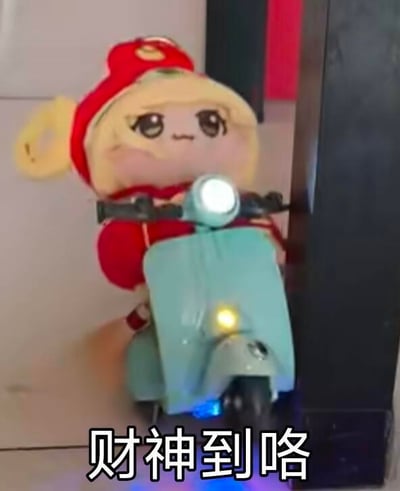 这是一张摆拍的趣味梗图，画面中是戴着红色财神风格帽子的《原神》芭芭拉衍生棉花娃娃，它坐在一辆带有灯光效果的浅蓝色玩具小摩托上，画面下方配有黑色文字“财神到咯”。用可爱的卡通玩偶形象结合迎财神的节日主题，营造出喜庆又诙谐的氛围，适合在春节、初五迎财神等节点用来传递祝福，没有双关、谐音梗等特殊含义。