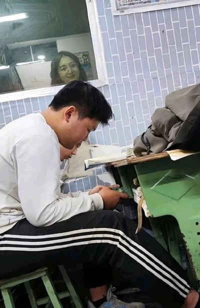 教室场景中，一名穿着白色长袖上衣和黑色条纹运动裤的男生坐在绿色凳子上，正低头专注于手中的物品。他面前是一张绿色的旧课桌，桌上放有一些杂物。背景是蓝白相间的瓷砖墙，墙上挂着一面镜子，镜中反射出一位面带微笑的女性（疑似老师）。整个画面呈现出校园日常的温馨氛围。