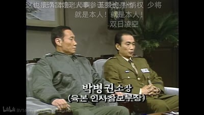 这是一段韩国军事人物访谈的视频截图，画面中右侧是陆军本部人事参谋部长朴炳权少将，他身着卡其色军装，双手交握置于腿上，神情严肃地看向斜下方；左侧是另一位身着军绿色便服的军人，侧头看向一旁，神情同样严肃。画面上方有B站用户添加的中文弹幕：“这也是陆军本部老人事参谋部长是朴炳权 少将 就是本人！就是本人！双日凌空”，画面下方有原视频的韩文标注“박병권 소장 (육본 인사참모부장)”，对应中文翻译为“朴炳权 少将（陆军本部人事参谋部长）”，整体场景为正式的访谈场合，氛围庄重严肃。
