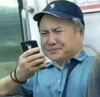 图片中是一位中年男子，他戴着蓝色帽子，穿着蓝色衬衫，正拿着一部黑色手机观看。男子眉头微皱，眼睛微眯，嘴角略带弧度，表情看起来有些疑惑又有点想笑。背景似乎是在公共交通工具内，能看到车窗和扶手。