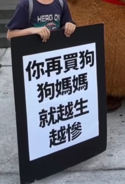 整张图为户外实拍的静态画面，一名穿着印有“HERO OX”字样深蓝色短袖、背着灰色双肩包的人士站在水泥地面上，双手扶着一块黑框白底的立牌，立牌上印有黑色繁体大字“你再買狗 狗媽媽 就越生 越慘”，内容直指购买繁殖犬会加剧母犬被迫繁殖的痛苦，以此呼吁大众选择领养代替购买宠物狗，减少犬只无序繁殖的情况。