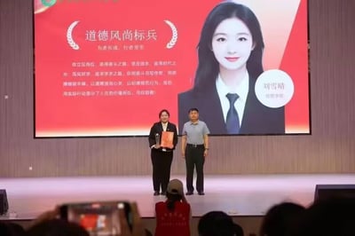 这是一场校园荣誉颁奖活动的现场实拍照片。舞台上左侧站着获奖的刘雪晴，她身着黑色正装，面带微笑，手持奖杯与红色荣誉证书；右侧是一位男性颁奖嘉宾，身着浅灰色上衣与深色长裤，站姿端正。背景的大型红色屏幕上展示着本次荣誉“道德风尚标兵”的主题，配有刘雪晴的正装照片、所属学院“经营学院”，以及一段详细的表彰文案，赞扬她立足岗位奋斗、笃实好学，以品德和行动彰显人生价值。台下有观众正用手机拍摄记录这一时刻，其中一位穿着红色志愿服的人员也在拍照，现场氛围正式庄重，充满荣誉感。