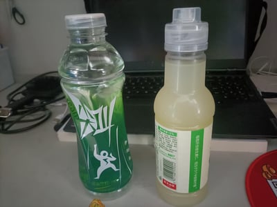 办公桌上并排放置着两瓶饮料，左侧是透明瓶身绿色标签的运动饮料，右侧是淡黄色液体的农夫山泉维生素饮料。背景中可见一台打开的笔记本电脑，桌面上还有一些电线等物品。