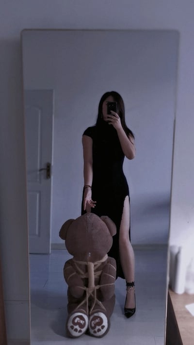 室内场景中，一名女子站在全身镜前自拍。她身着黑色无袖长裙，裙摆侧面有高开叉设计，脚踩黑色高跟鞋，右手举着手机拍摄镜中的自己。镜前地面上坐着一个棕色泰迪熊玩偶，其腿部被绳子捆绑。房间整体为简约的白色调，背景中可见一扇关闭的白色房门，光线柔和偏冷，营造出略带神秘感的氛围。