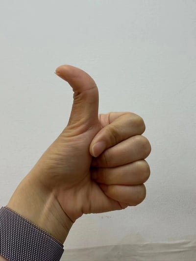 一张特写照片，展示了一只真实人物的手做出竖大拇指的手势。手部皮肤纹理清晰可见，手腕处露出深色蓝色格子图案的衣袖，背景为纯白色墙面，整体构图简洁，突出手势本身，传递出认可或赞赏的积极信号。
