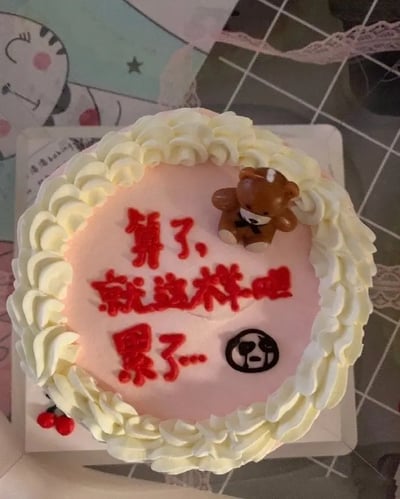 这是一款装饰有白色奶油花边和小熊玩偶的粉色蛋糕，蛋糕中央用红色奶油写着“算了，就这样吧，累了...”，并配有一个无奈/疲惫的表情符号。这句写在蛋糕上的话常常用来表达对某事已经疲惫不堪、随遇而安的心态，自带一种自嘲和无奈的幽默感，很适合送给正在经历压力、感到疲倦的朋友，或者用来调侃自己。文字和表情形成了明显的自嘲和释然氛围，带有轻微的摆烂意味。蛋糕整体风格较为少女且治愈，但搭配的文字内容形成反差萌。没有明显的双关或谐音梗。