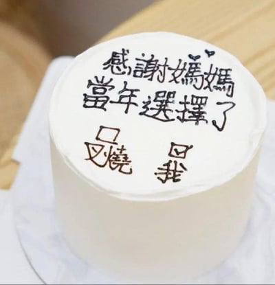 一张圆形白色奶油蛋糕的特写照片，蛋糕顶部用黑色巧克力酱手写着“感谢妈妈当年选择了我”，旁边点缀着两个小爱心图案。蛋糕放置在白色纸托上，背景是浅色木质桌面，整体氛围温馨又带点幽默，表达了对母亲的感恩之情。