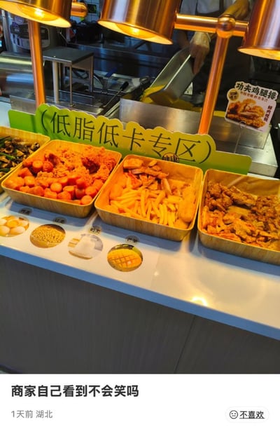 这是一张网友实拍的线下餐饮门店餐台图搭配吐槽文字的社交媒体内容，画面上方的餐台摆放着盛有炸香肠、炸鸡块、薯条、炸鸡排等高热量油炸食物的餐盒，餐台后方立有写着“（低脂低卡专区）”的绿色标识牌，旁边还有“功夫鸡腿排”的产品宣传卡，背景可见门店工作人员正在操作厨具；图片下方配有发布者的吐槽文字“商家自己看到不会笑吗”，标注了发布地点为湖北、发布时间为1天前，内容讽刺商家虚假宣传，将明显高油高热量的油炸食品区域标榜为低脂低卡区，反差感十足充满喜剧效果。