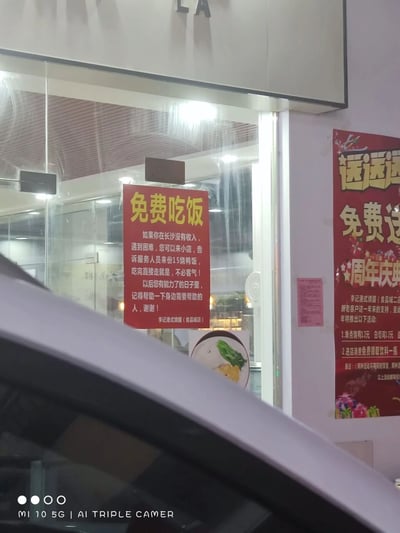 这是一张用MI 10 5G手机拍摄的长沙李记港式烧腊（食品城店）门口的照片。玻璃门上张贴着一张红底黄字的“免费吃饭”爱心告示，告示内容为：为在长沙没有收入、遭遇困难的人群提供免费的15烧鸭饭，无需客气可直接用餐离开，同时呼吁受助者在有能力后去帮助身边需要帮助的人，传递善意。在店铺右侧的墙面上，还贴着该店的周年庆促销海报，海报以红色为主色调，写有“送送送 免费送”等促销字样，列出了单点烧鸭优惠、进店领饮料等活动。画面左下角能看到车辆的部分车身，整体场景传递出商家的公益善意与促销信息。