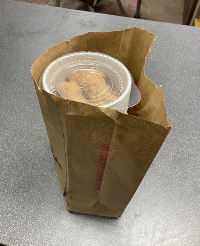 一张近距离拍摄的食物照片，展示了一个放在灰色桌面上的牛皮纸袋，袋中装着一个带透明塑料盖的圆形容器，容器内可以看到一块饼干。背景中隐约可见桌椅，整体呈现出日常用餐场景的随意感。