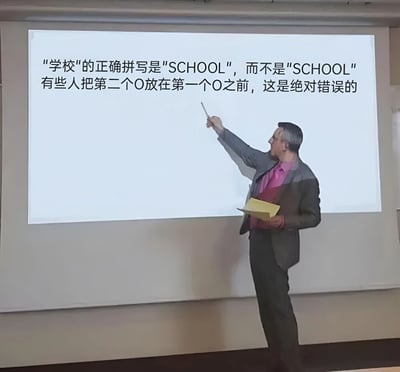 一位穿着西装的男子手持黄色文件夹，正用指示棒指向投影屏幕。屏幕上用中文写着："学校的正确拼写是'SCHOOL'，而不是'SCHOOL' 有些人把第二个O放在第一个O之前，这是绝对错误的"。实际上两句中的'SCHOOL'拼写完全相同，这种一本正经地纠正不存在错误的行为形成了滑稽的幽默效果。