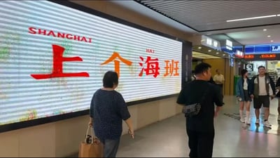 这张图片拍摄于上海某地铁站的公共通行通道内，画面左侧设置了一块大型电子宣传屏，屏幕原本应展示城市宣传内容“上海 SHANGHAI”，但因显示故障巧合形成玩梗效果，文字被拆分组合成了“上个海班”的字样，其中“上”和“海”为红色字体，“个”和“班”为橙色字体，“海”字上方还保留了原有的拼音标注“HAI”。通道内有数名行人通行，一名提着纸袋的中老年女性正看向屏幕，另有多名乘客或行走或驻足，背景可见地铁站的出口标识与周边商铺招牌。该内容玩“去上海上班”的谐音梗，用来调侃在上海打拼的打工人的工作状态，引发了网友的共鸣与传播。