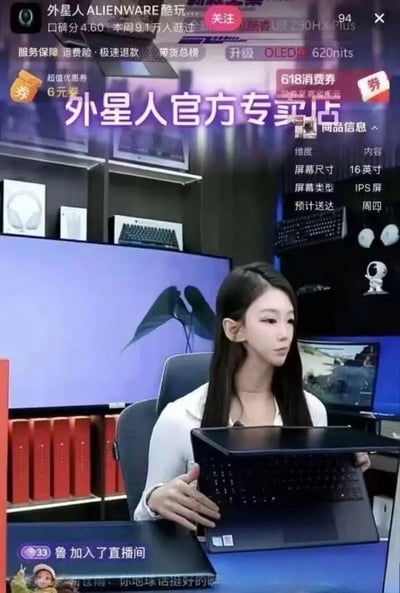 这是一张电商直播截图，为外星人ALIENWARE电脑官方专卖店的直播场景，画面中的女主播有着外形尖锐细长的精灵耳，恰好和其售卖的“外星人”品牌产品形成呼应，产生趣味梗。画面上层叠加了直播间的各类UI元素：左上角显示店铺评分、浏览人数，还有6元优惠券、618消费券等福利提示；中部大字标注“外星人官方专卖店”；右上角列出在售笔记本的参数：16英寸IPS屏，预计周四送达；底部显示有观众进入直播间的提示，还有观众调侃的弹幕“你地球话挺好的啊”。背景陈列着耳机、键盘等数码周边产品，主播正手持外星人笔记本电脑进行展示。