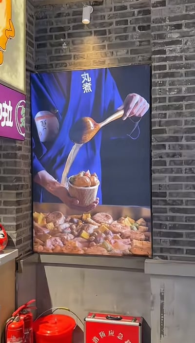 这是一张拍摄于店内的宣传海报照片，画面中有一位穿着蓝色制服的人正用勺子把高汤或汤汁倒进装满火锅关东煮食材的碗里。背景是各色关东煮食材，人物制服胸前有“丸煮”字样，极具日式风格，营造出仪式感，食欲满分。下方的实体环境能看到灭火器和消防箱，突出了店铺内实际拍摄的生活氛围。
没有明显的双关、谐音梗或暗示。