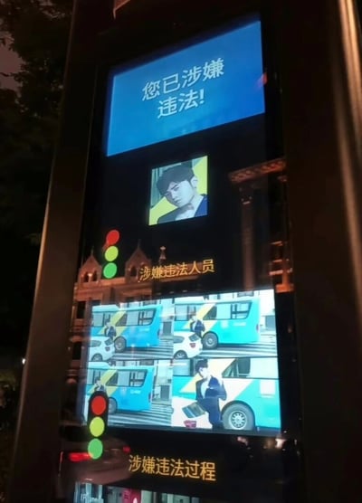 这是一张经过P图改编的恶搞类图片，模拟了城市街头的交通违法曝光电子屏界面。界面顶部蓝色区域醒目显示“您已涉嫌违法！”的提示语，中间区域标注“涉嫌违法人员”，使用了歌手周杰伦的肖像；下方“涉嫌违法过程”区域则截取了周杰伦某支MV中的画面，将其伪装成闯红灯的交通违法场景。整体内容是将明星的影视素材与真实的交通曝光系统界面相结合，通过错位的场景营造出荒诞搞笑的趣味效果，属于网友创作的明星相关趣味梗图。
