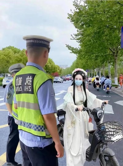 这张图片展现了一名穿着古风汉服和花环头饰的女生，骑着电动车在马路上被交警拦下。现代交警与古风装扮女生形成鲜明反差，很有趣味性和梗感。没有明显的双关或谐音梗，但画面夸张戏剧，适合玩穿越和仪式感等话题。