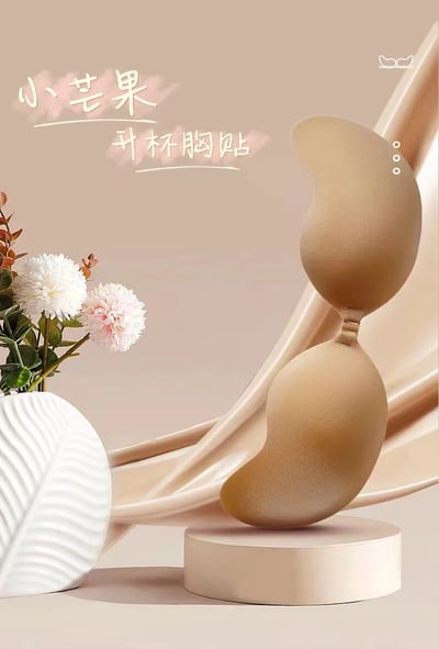 这是一张产品宣传图片，展示了一款名为"小芒果升杯胸贴"的女性用品。图片主体是一个米色的芒果形状胸贴，放置在一个圆形的展示台上。胸贴旁边有一个白色的花瓶，里面插着白色和粉色的花朵。背景是柔和的米色，有流动的布料元素增添优雅感。整体构图精致，色调统一，营造出高端、优雅的产品形象。