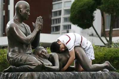 这是一张户外拍摄的趣味照片，画面中有一组青铜材质的公共艺术雕像：一位光头和尚盘腿端坐，双手合十呈祈祷姿态，神情平静；旁边是一位侧卧在地的女性雕像，神态放松。一个穿着白红拼接短袖与花短裤的男孩，趴在女性雕像的背部，身体的位置和姿势与雕像形成了巧妙的视觉错位，看起来仿佛男孩正在和女性雕像进行亲密互动，而一旁的和尚雕像则如同在注视这一幕，整体场景充满滑稽感，是游客利用雕像进行的创意摆拍，制造出了充满趣味的巧合画面。