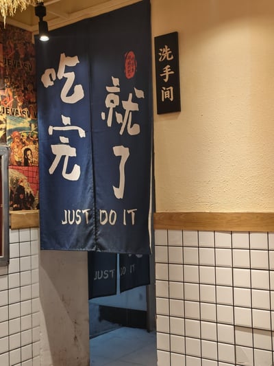 这是一家餐厅洗手间入口的照片，入口处挂着蓝色门帘，上面写着“吃完就了”，英文则为“JUST DO IT”。一旁墙上有“洗手间”标识。中文和英文结合，幽默地暗示“吃完饭马上去厕所”，结合了Nike的著名广告语、双关和谐音梗，形成梗图。整体风格搞怪轻松，适合转发吐槽。
