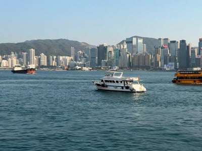 这张图片展示了香港维多利亚港的壮丽景色。画面中可以看到平静的蓝色海面上有三艘船：中央是一艘白色游艇，右侧是一艘黄色双层船，左侧远处有一艘深色货船。背景是香港标志性的城市天际线，由众多高楼大厦组成，其中可以清晰看到带有"Panasonic"标志的建筑。城市后方是连绵的绿色山脉，天空呈现出清澈的淡蓝色，整体画面给人一种宁静而繁华的感觉。
