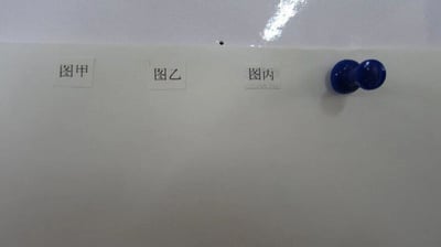 一张白板的照片，上面贴有三个标签，分别是左侧的'图甲'、中间的'图乙'和右侧的'图丙'。白板右上角有一个蓝色的图钉，整体背景为纯白色，表面有轻微反光。
