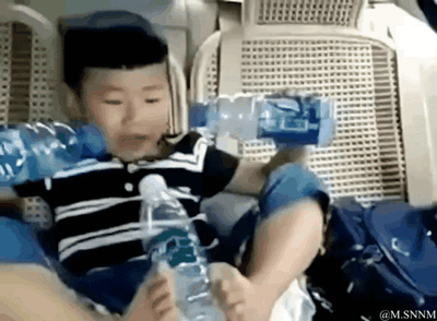 这是一段搞笑的动态GIF，画面里的小男孩坐在汽车座椅上，通过巧妙的视觉错位拍摄手法，让他的脚和手分别握持矿泉水瓶，从镜头角度看仿佛小男孩长了四只手，同时玩转三个矿泉水瓶，画面充满趣味，利用视觉错觉制造出滑稽又欢乐的效果。