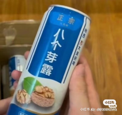 这是一只手拿着一罐山寨饮料，罐身主打蓝白配色设计，顶部标有“正宗”字样，中间是醒目的“八个芽露”大字，侧面写着“喝西大大清好喝”，罐身下方还有核桃与绿叶的图案，明显是模仿知名核桃露饮料“六个核桃”的山寨产品。
这是典型的蹭热度仿冒饮品，通过模仿知名品牌的包装风格与命名逻辑，用“八个芽露”的名称制造出滑稽的反差感，玩了名称模仿的搞笑梗。