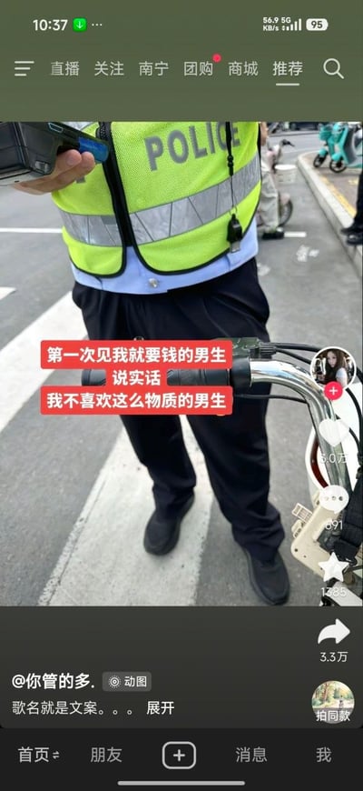 这是一张抖音平台的短视频截图，场景为城市马路的斑马线区域，一名身着印有“POLICE”字样荧光绿反光执勤背心的交通警察站在画面中央，手持设备正对拍摄者，拍摄者当时骑坐在电动车上，画面可看到电动车车把，交警疑似正在处理拍摄者的交通违法行为、准备开具罚单。画面中叠加了红色背景的白色调侃文案，套用吐槽交往对象物质的常用话术戏说被交警罚款的场景，形成强烈反差笑点。画面右侧显示抖音的点赞、评论、收藏、转发交互按钮，底部显示发布者账号为@你管的多.，附带文案“歌名就是文案。。。”，顶部是抖音的导航栏，当前定位城市为南宁。