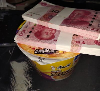 这是一张实拍的单图，深色桌面上放着一桶康师傅金汤系列桶装方便面，桶盖上摞放着两捆用封条捆好的人民币，下方一捆为一百元面额人民币且票面倒置摆放，上方一捆为十元面额人民币，整体画面制造出一种反差趣味，以金钱搭配平价泡面的组合形成调侃效果，带有趣味炫富的反差感。