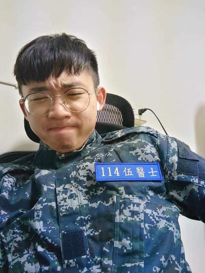 图中是一位身着带有「114伍醫士」标识的数码迷彩服的男生，他瘫坐在办公椅上，皱起眉头、挤着双眼，嘴角向下撇，露出委屈又崩溃的表情，看起来是在宣泄服勤工作带来的疲惫与无奈。