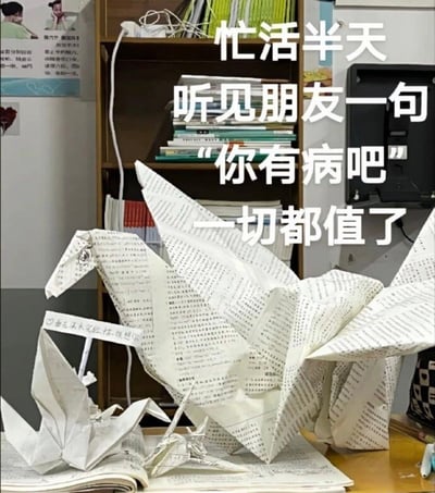 图片展示了一个用写满文字的书页折成的大型纸马和几只纸鹤，放置在书桌上。背景是一个堆满书籍的书架，墙上贴有一些海报和纸张。图片上方有醒目的白色文字："忙活半天 听见朋友一句'你有病吧' 一切都值了"，表达了一种自嘲式的幽默——花费大量时间创作的手工艺术品被朋友调侃，但创作者依然觉得值得。前景中还有一个小标签写着："艺术来源于生活 你想你"。整体场景呈现出一种文艺又带点搞笑的氛围。