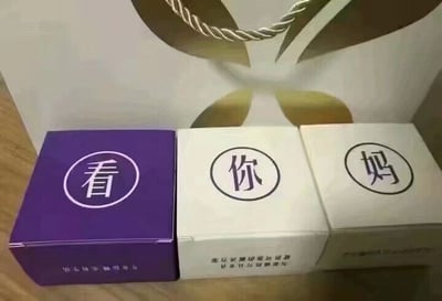这是一张静态单图，画面展示了三个方形包装盒，从左到右依次为紫色、白色、白色包装盒，每个盒子正面的圆形边框内分别印有“看”“你”“妈”三个汉字，将三个盒子并排摆放后，文字连起来是带有调侃、冒犯意味的口语化表述。盒子底部印有倒置的小字，紫色盒子底部是“为你精选品质生活”，白色盒子底部是“为您提供专业可靠的解决方案”，背景是一个带有金色装饰图案和绳状提手的白色手提袋。这张图通过包装盒上的文字排列制造出趣味调侃的效果，属于利用文字拼接创作的整蛊梗图。