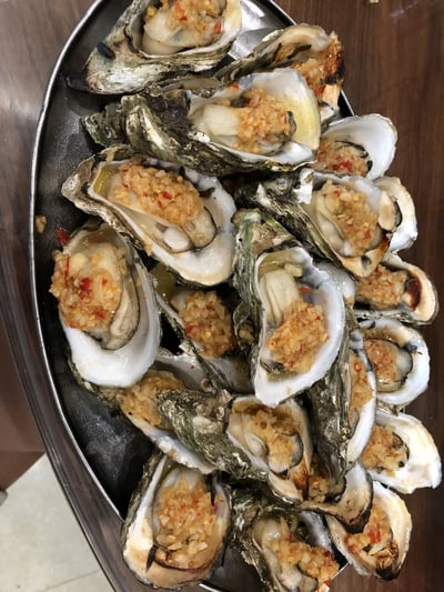 一张俯拍视角的美食照片，展示了满满一盘蒜蓉烤生蚝。生蚝壳打开后露出饱满的蚝肉，上面覆盖着金黄色的蒜蓉和红色辣椒碎，排列在椭圆形金属托盘上，背景为木质桌面。生蚝个头大小不一，部分边缘有轻微烤焦痕迹，整体色泽诱人，呈现出刚出炉的鲜香质感。