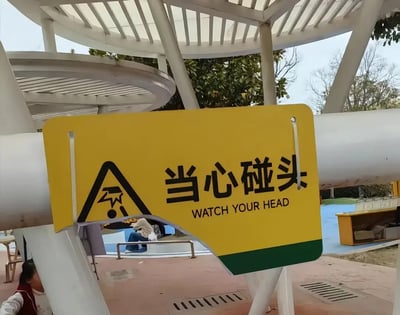 这是一张户外实拍静态图，画面主体是一块安装在白色管状公共设施上的黄色警示标识，标识左侧印有碰头警示图案，右侧印有中文“当心碰头”及对应英文“WATCH YOUR HEAD”的提示内容。但该警示牌本身安装位置极低，路过的行人首先就会被这块警示牌碰到头部，和提示内容形成了极具讽刺感的反差笑点。背景可见户外白色廊架结构、绿植，以及类似公园或休闲园区的公共场地，画面左下角和背景处还能看到零星的行人。