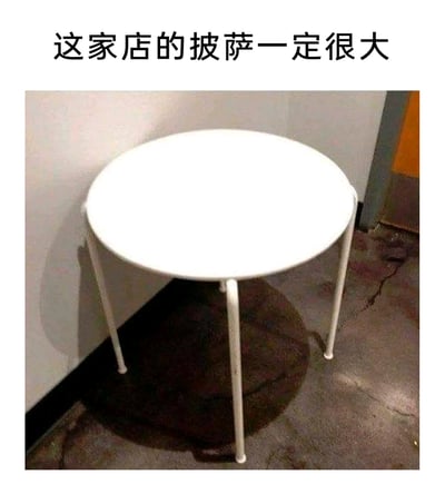 这是一张利用视觉错位效果制造笑点的梗图。画面中的白色圆形小桌倚靠在墙角，墙角的阴影恰好与其中一条桌腿的影子重合，使得桌子看起来像是被切去了三分之二的披萨。搭配上方的文字“这家店的披萨一定很大”，通过这个巧妙的视觉错觉，调侃原本的披萨尺寸巨大，仅剩下的“一角”就有桌子这么大，以此制造出荒诞搞笑的趣味效果。